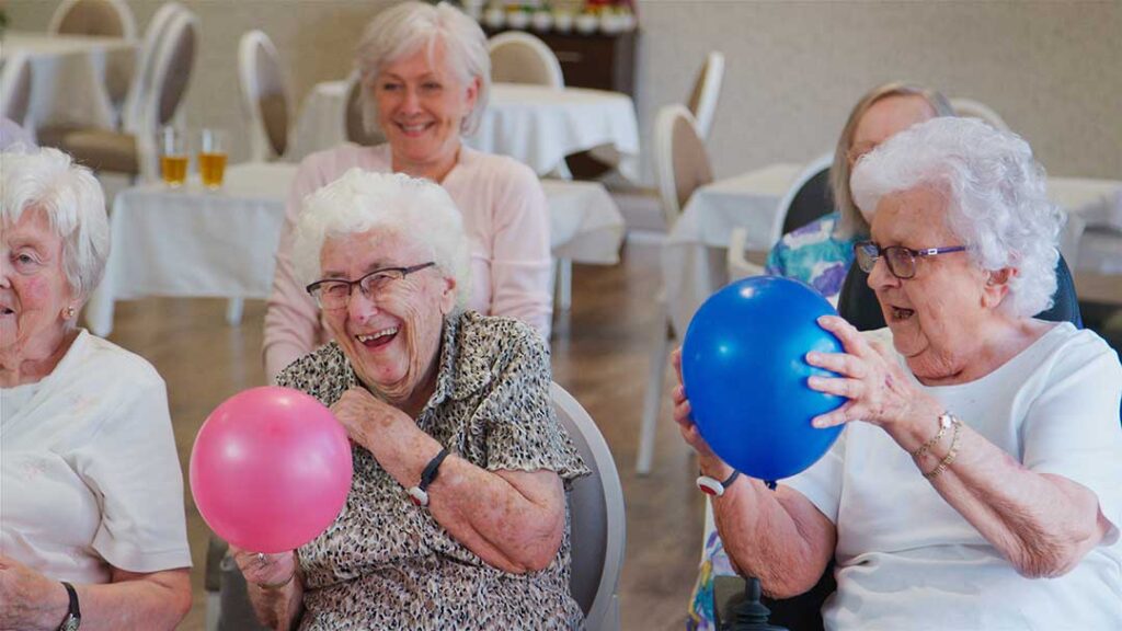 Two older adults sitting together in a comfortable common area while a caregiver assists in the background.