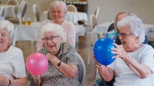 Two older adults sitting together in a comfortable common area while a caregiver assists in the background.