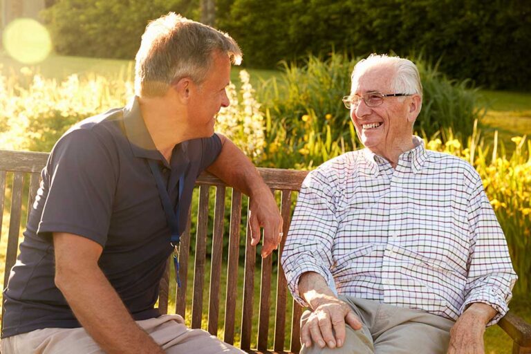 A senior resident and caregiver talking together at a dining table in a bright, comfortable shared living space.